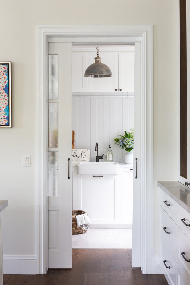 Inspiration for a mid-sized transitional galley utility room in Sydney with a farmhouse sink, shaker cabinets, white cabinets, marble benchtops, grey splashback, shiplap splashback, white walls, porcelain floors, a stacked washer and dryer, grey floor, grey benchtop, recessed and panelled walls.