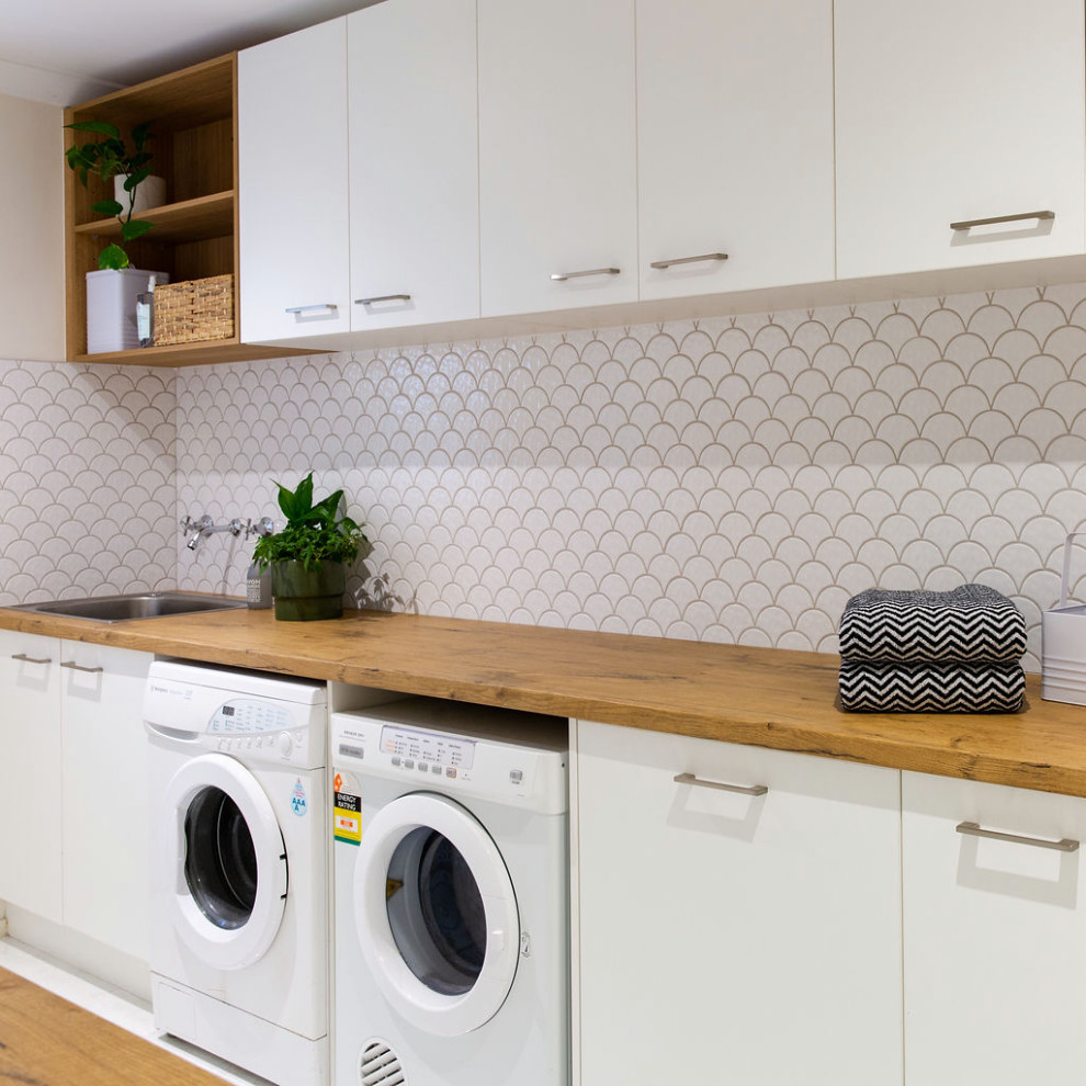 This is an example of a mid-sized modern galley dedicated laundry room in Melbourne with a drop-in sink, white cabinets, laminate benchtops, white splashback, porcelain splashback, white walls, medium hardwood floors and a side-by-side washer and dryer.