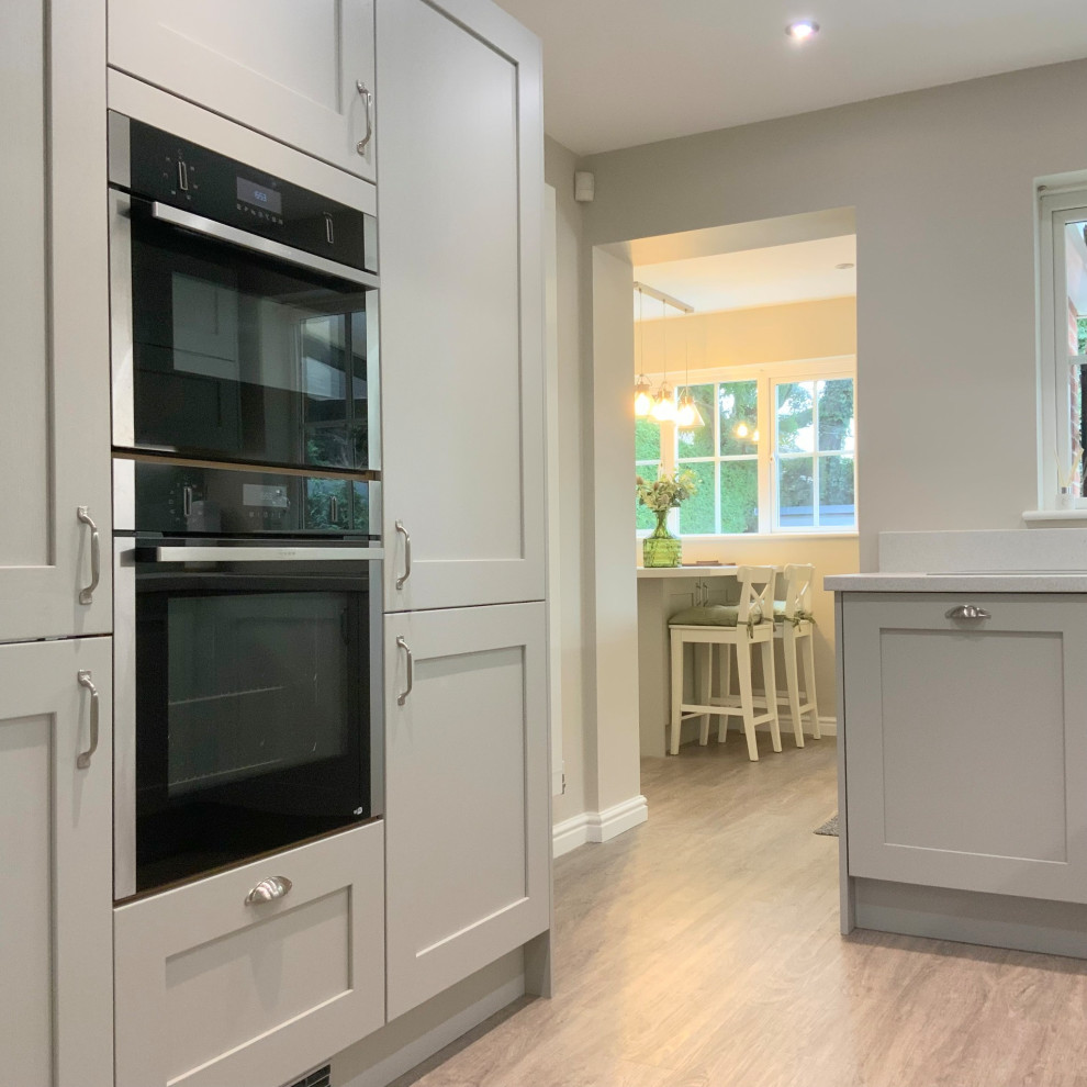 This is an example of a medium sized contemporary grey and white l-shaped kitchen/diner in Other with shaker cabinets, grey cabinets, laminate countertops, grey splashback, glass sheet splashback, integrated appliances, vinyl flooring, a breakfast bar, brown floors and white worktops.