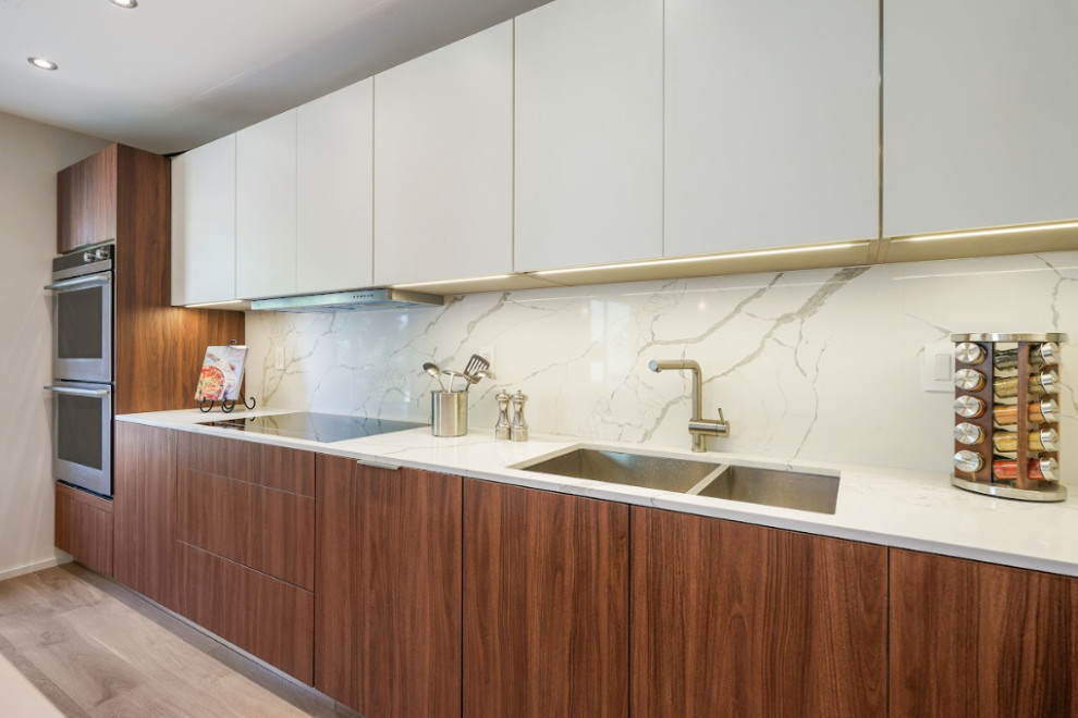 This is an example of a medium sized contemporary single-wall open plan kitchen in Los Angeles with a submerged sink, flat-panel cabinets, marble worktops, white splashback, marble splashback, stainless steel appliances, light hardwood flooring, an island, brown floors, white worktops and white cabinets.