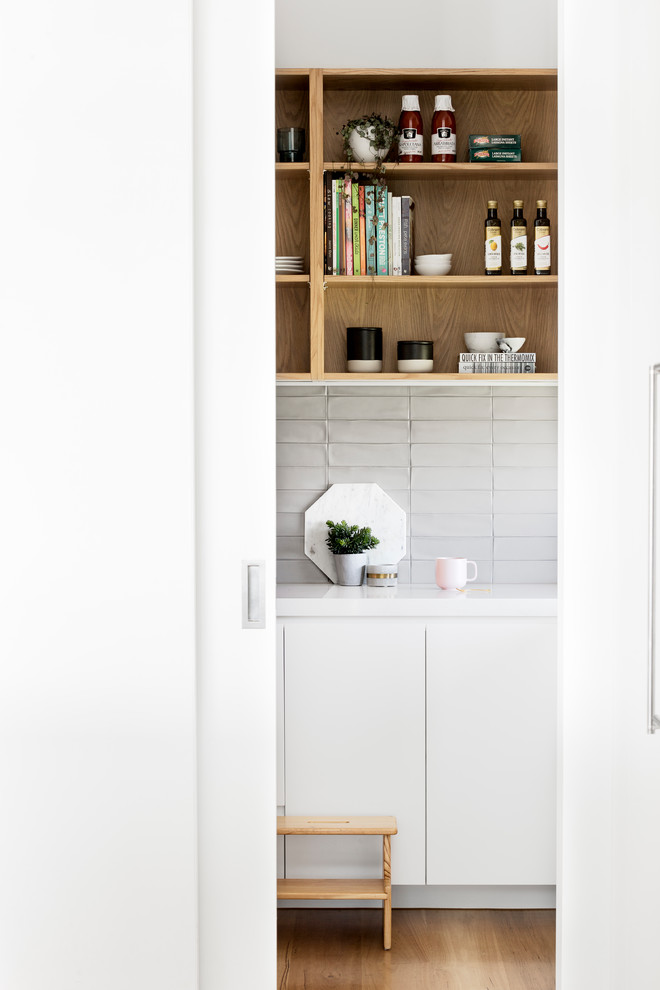 This is an example of a contemporary galley kitchen pantry in Melbourne with flat-panel cabinets, white cabinets, quartz benchtops, grey splashback, ceramic splashback, medium hardwood floors, brown floor and white benchtop.