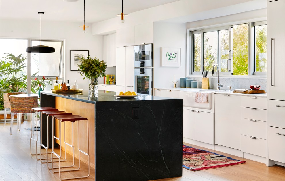 This is an example of a midcentury single-wall eat-in kitchen in Los Angeles with a farmhouse sink, flat-panel cabinets, white cabinets, panelled appliances, light hardwood floors and with island.