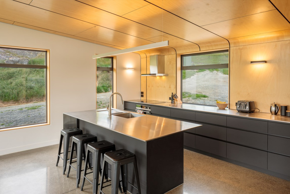 This is an example of a mid-sized contemporary l-shaped eat-in kitchen in Christchurch with a double-bowl sink, flat-panel cabinets, black cabinets, stainless steel benchtops, multi-coloured splashback, timber splashback, stainless steel appliances, concrete floors, with island, grey floor and grey benchtop.