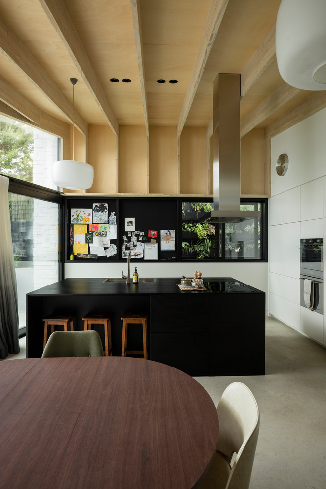 This is an example of a mid-sized contemporary galley eat-in kitchen in Auckland with a drop-in sink, flat-panel cabinets, white cabinets, stainless steel appliances, concrete floors, with island, grey floor and wood benchtops.