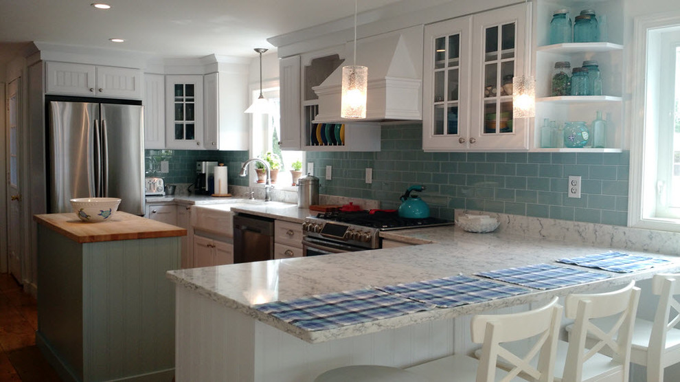 This is an example of a large beach style u-shaped eat-in kitchen in Portland Maine with a farmhouse sink, louvered cabinets, white cabinets, marble benchtops, blue splashback, subway tile splashback, stainless steel appliances, medium hardwood floors and with island.