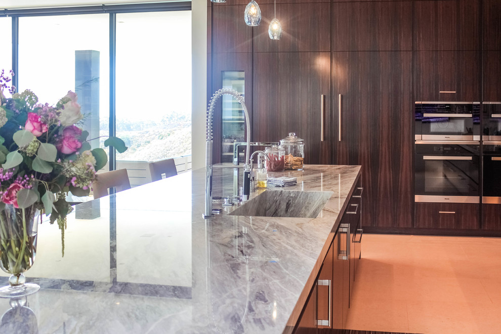 Large modern l-shaped eat-in kitchen in Los Angeles with beige floor, an integrated sink, flat-panel cabinets, dark wood cabinets, granite benchtops, grey splashback, stone slab splashback, stainless steel appliances, travertine floors, with island and grey benchtop.