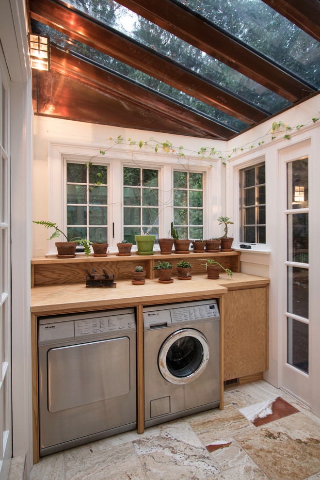 This is an example of a small traditional single-wall utility room in New York with flat-panel cabinets, light wood cabinets, wood benchtops, white walls, travertine floors, a side-by-side washer and dryer and beige benchtop.