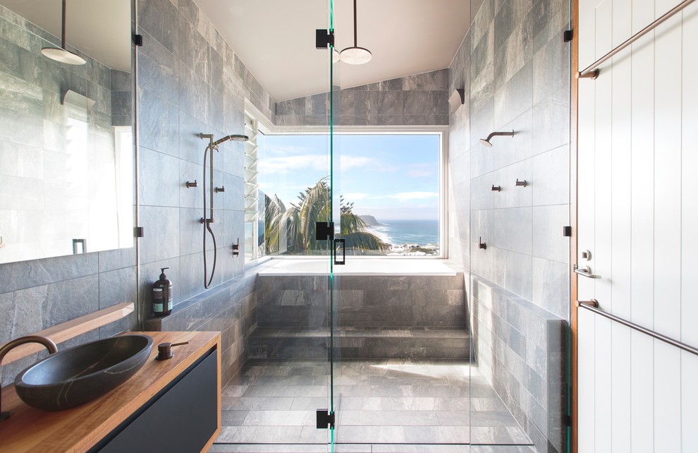 This is an example of a mid-sized beach style master bathroom in Newcastle - Maitland with a drop-in tub, a double shower, gray tile, ceramic tile, grey walls, ceramic floors, wood benchtops, grey floor, flat-panel cabinets, black cabinets, a vessel sink, a hinged shower door and brown benchtops.