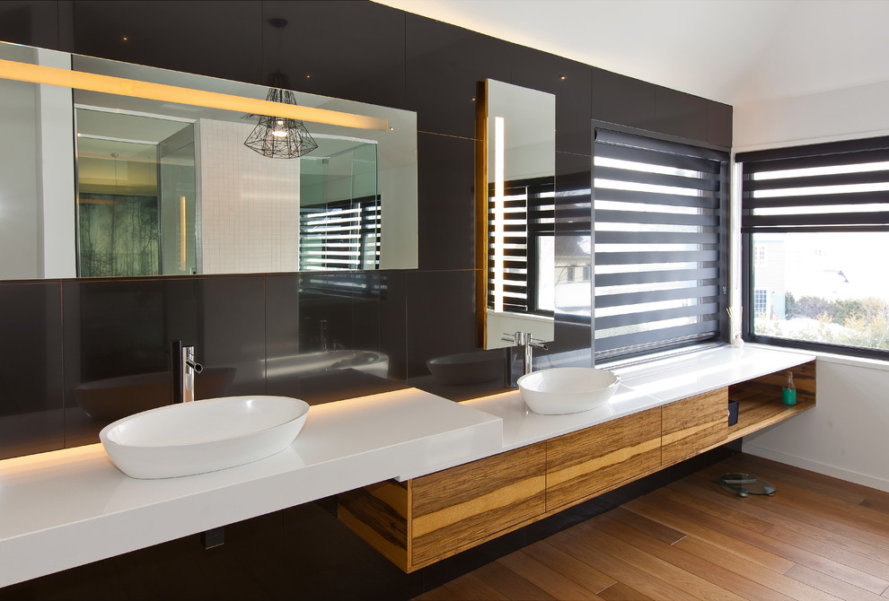 Large modern master bathroom in Montreal with flat-panel cabinets, medium wood cabinets, a freestanding tub, black tile, ceramic tile, white walls, light hardwood floors, a vessel sink, solid surface benchtops and beige floor.
