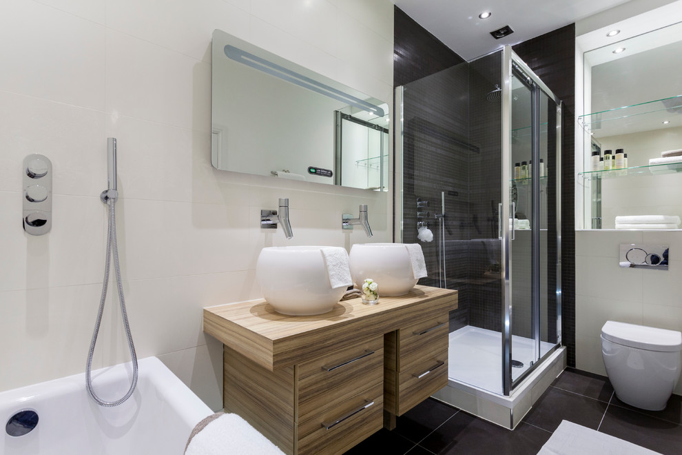 Photo of a contemporary bathroom in London with a vessel sink, flat-panel cabinets, light wood cabinets, wood benchtops, a corner shower, a wall-mount toilet, black tile and white walls.