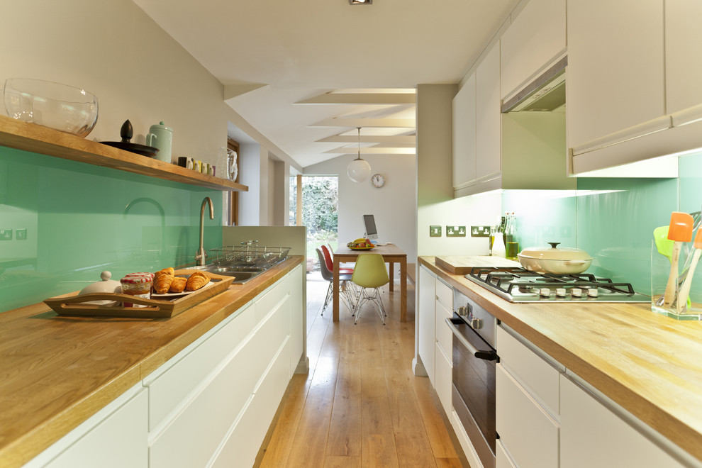 Inspiration for a midcentury galley separate kitchen in Other with stainless steel appliances, wood benchtops, a drop-in sink, flat-panel cabinets, white cabinets and glass sheet splashback.