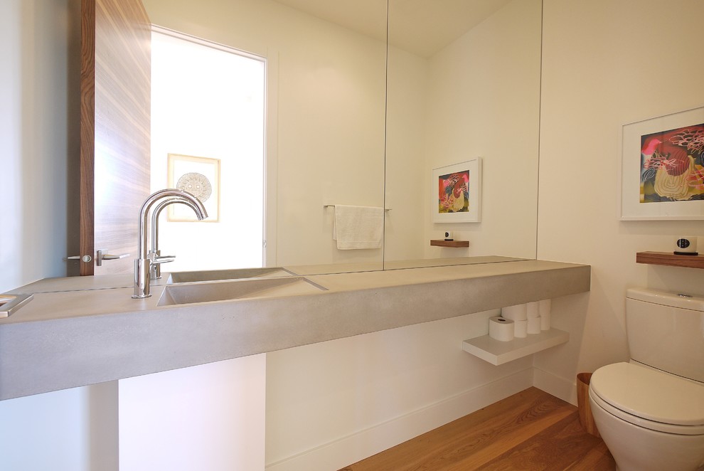 This is an example of a mid-sized modern powder room in Calgary with white walls, an integrated sink, concrete benchtops, a two-piece toilet, medium hardwood floors, white cabinets and grey benchtops.