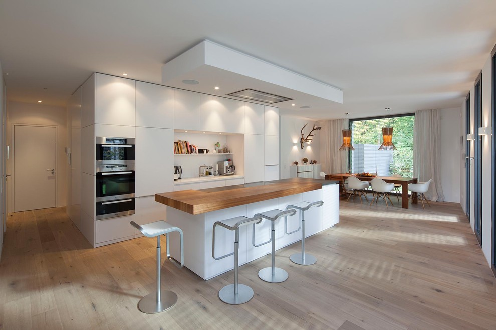 Photo of a large contemporary single-wall open plan kitchen in Cologne with medium hardwood floors, flat-panel cabinets, white cabinets, panelled appliances, with island, an integrated sink and white splashback.
