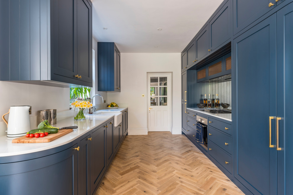 Inspiration for a large transitional galley eat-in kitchen in London with a farmhouse sink, shaker cabinets, blue cabinets, solid surface benchtops, mirror splashback, stainless steel appliances, no island and white benchtop.