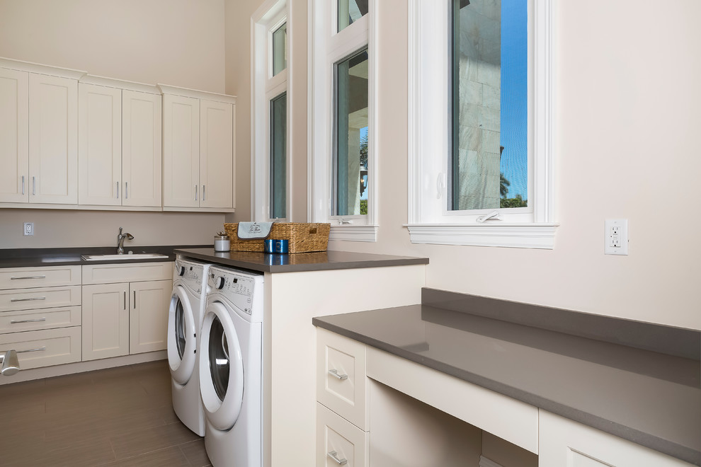 Photo of a mid-sized tropical l-shaped utility room in Miami with recessed-panel cabinets, white cabinets, quartz benchtops, a side-by-side washer and dryer, a drop-in sink, white walls and porcelain floors.