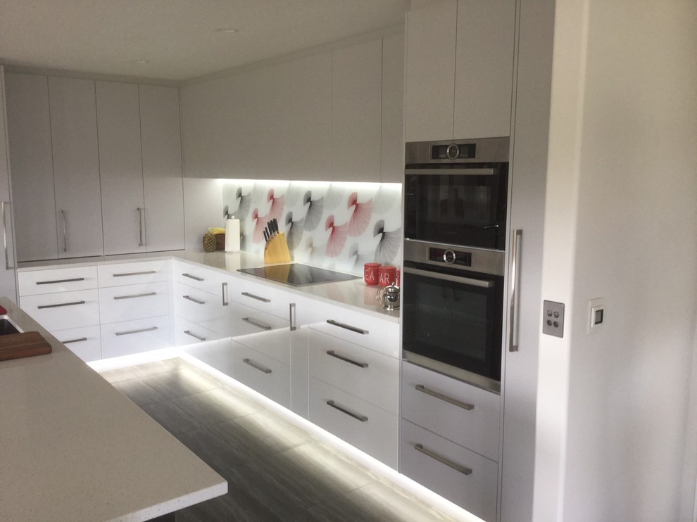 Photo of a large contemporary l-shaped open plan kitchen in Christchurch with a single-bowl sink, flat-panel cabinets, white cabinets, quartz benchtops, multi-coloured splashback, glass sheet splashback, stainless steel appliances, vinyl floors and with island.
