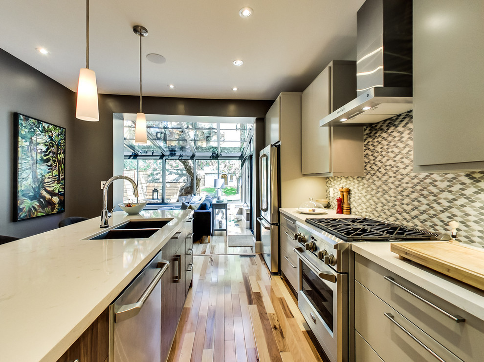 Inspiration for a mid-sized transitional galley light wood floor and multicolored floor kitchen remodel in Toronto with a double-bowl sink, flat-panel cabinets, beige cabinets, quartz countertops, gray backsplash, ceramic backsplash, stainless steel appliances and an island