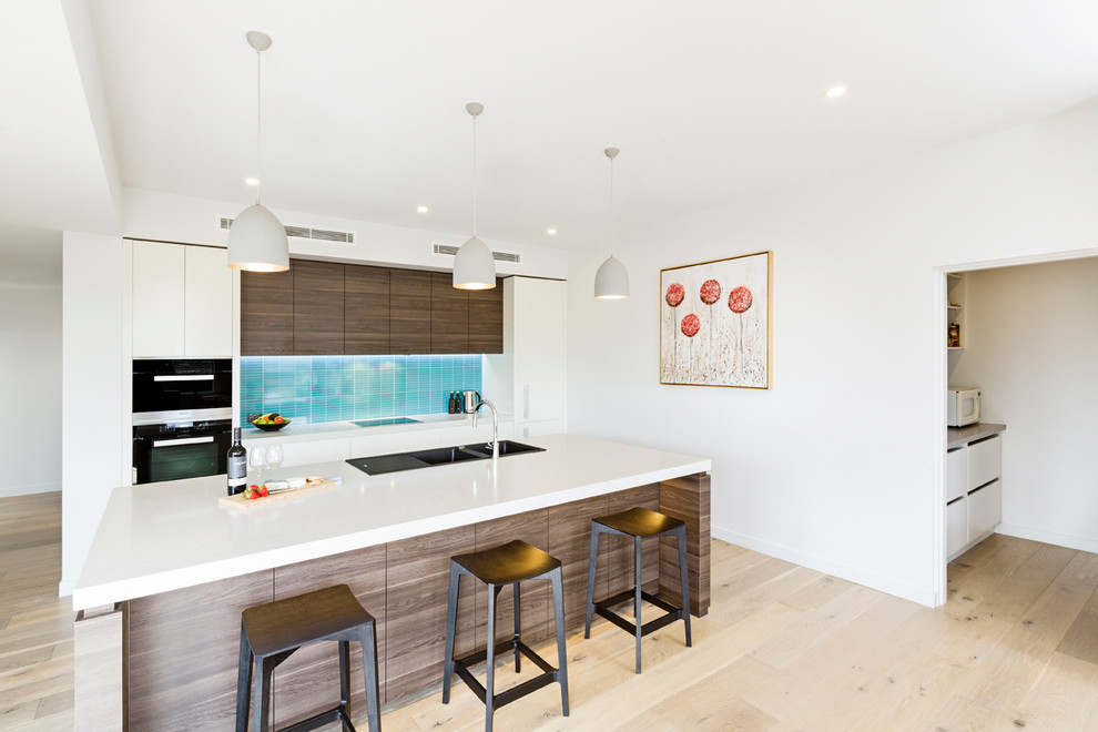 This is an example of a mid-sized contemporary single-wall eat-in kitchen in Melbourne with a double-bowl sink, flat-panel cabinets, white cabinets, quartz benchtops, blue splashback, ceramic splashback, black appliances, light hardwood floors, with island and beige floor.