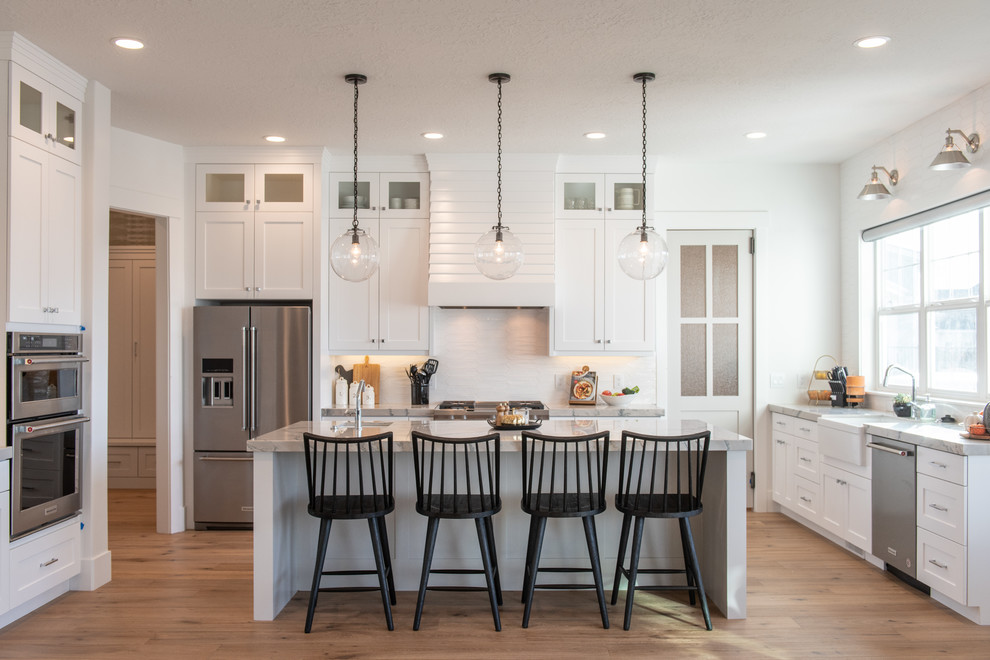 This is an example of a large transitional u-shaped separate kitchen in Salt Lake City with a farmhouse sink, shaker cabinets, white cabinets, marble benchtops, white splashback, stainless steel appliances, light hardwood floors, with island, beige floor, grey benchtop and porcelain splashback.