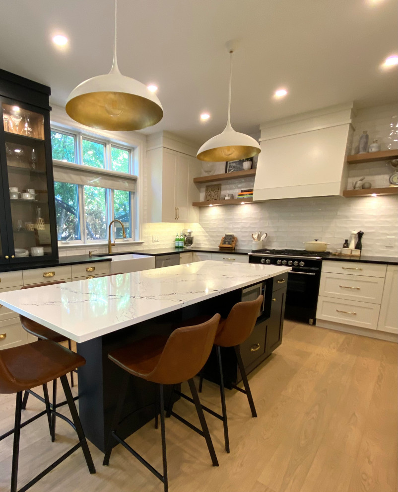 Example of a mid-sized transitional l-shaped light wood floor open concept kitchen design in Toronto with a farmhouse sink, shaker cabinets, white cabinets, quartz countertops, white backsplash, porcelain backsplash, an island and black countertops