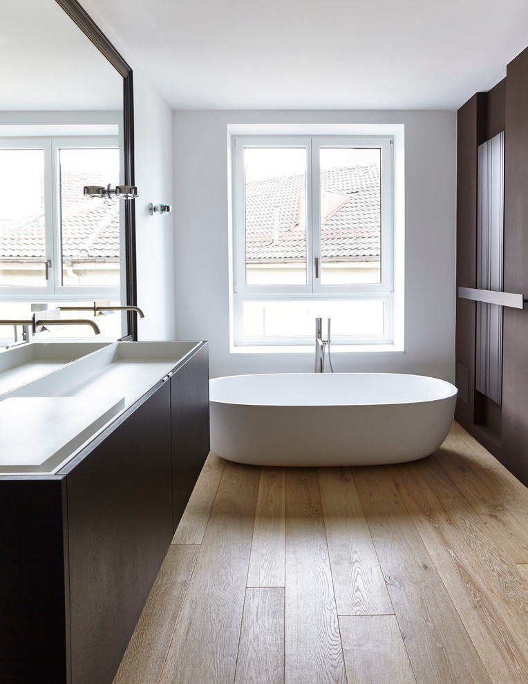 Design ideas for a mid-sized modern master bathroom in Munich with flat-panel cabinets, dark wood cabinets, a freestanding tub, light hardwood floors, white walls, solid surface benchtops and a trough sink.