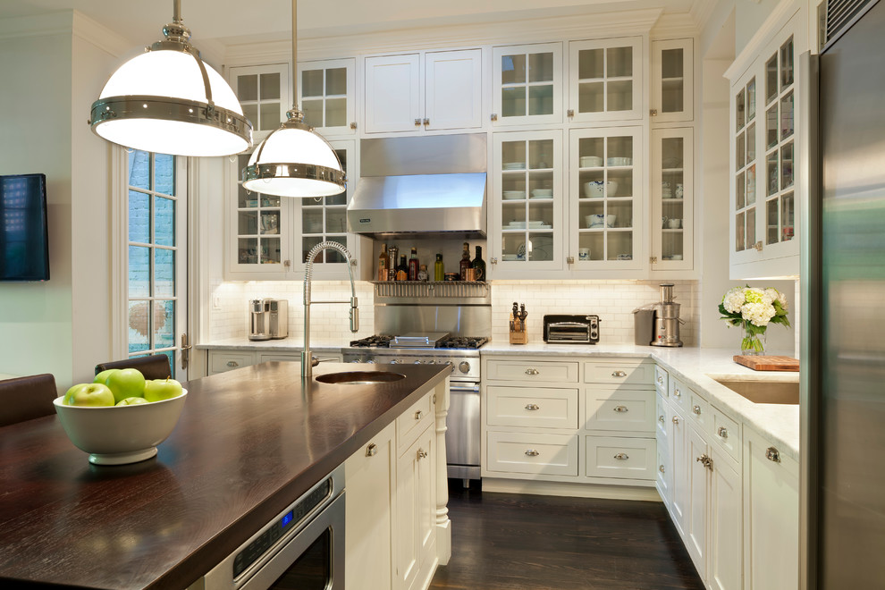 This is an example of a country l-shaped eat-in kitchen in DC Metro with glass-front cabinets, white cabinets, marble benchtops, white splashback, stainless steel appliances, dark hardwood floors, with island and subway tile splashback.