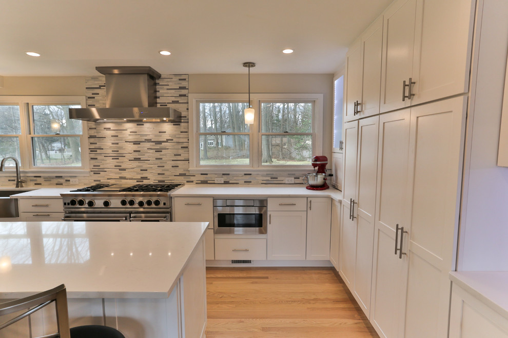 Photo of a large transitional u-shaped eat-in kitchen in Philadelphia with a farmhouse sink, recessed-panel cabinets, white cabinets, quartz benchtops, grey splashback, matchstick tile splashback, stainless steel appliances, light hardwood floors, with island, beige floor and white benchtop.