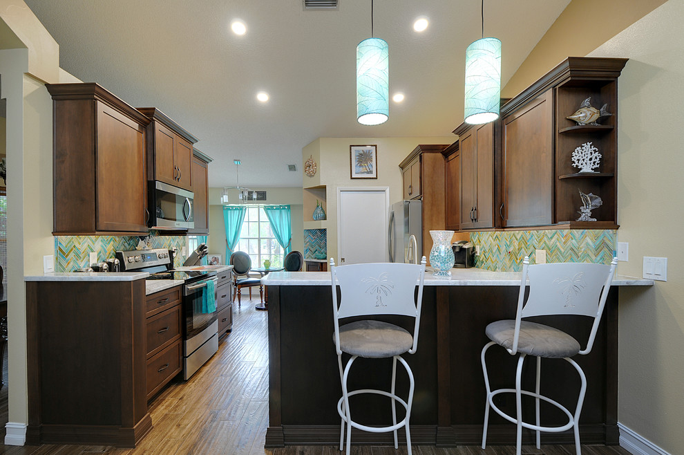This is an example of a mid-sized tropical u-shaped eat-in kitchen in Tampa with an undermount sink, shaker cabinets, medium wood cabinets, granite benchtops, brown splashback, glass tile splashback, stainless steel appliances, porcelain floors and a peninsula.