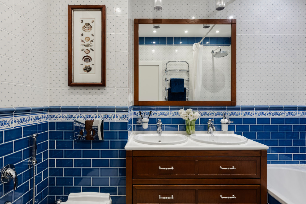 Photo of a mid-sized traditional bathroom in Saint Petersburg with brown cabinets, an undermount tub, a wall-mount toilet, blue tile, ceramic tile, white walls, ceramic floors, a drop-in sink, engineered quartz benchtops, white benchtops, a double vanity, a freestanding vanity and shaker cabinets.