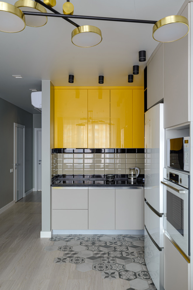 This is an example of a contemporary l-shaped kitchen in Novosibirsk with flat-panel cabinets, yellow cabinets, beige splashback, white appliances, light hardwood floors, no island, beige floor, black benchtop and subway tile splashback.