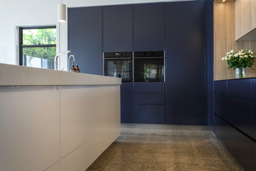 This is an example of a large contemporary l-shaped kitchen pantry in Brisbane with a double-bowl sink, flat-panel cabinets, light wood cabinets, quartz benchtops, mirror splashback, black appliances, concrete floors, with island, multi-coloured floor and white benchtop.