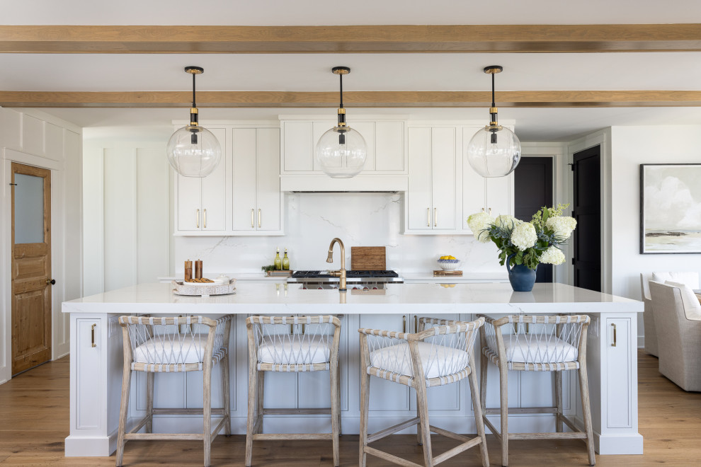 Open concept kitchen - coastal galley medium tone wood floor, brown floor and exposed beam open concept kitchen idea with a farmhouse sink, shaker cabinets, white cabinets, white backsplash, stone slab backsplash, stainless steel appliances, an island and white countertops