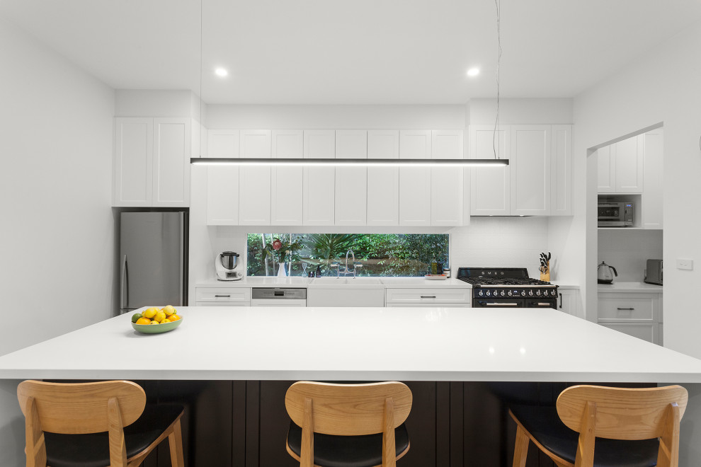 Inspiration for a mid-sized contemporary single-wall open plan kitchen in Melbourne with a farmhouse sink, shaker cabinets, white cabinets, quartz benchtops, white splashback, window splashback, black appliances, light hardwood floors, with island, beige floor and white benchtop.
