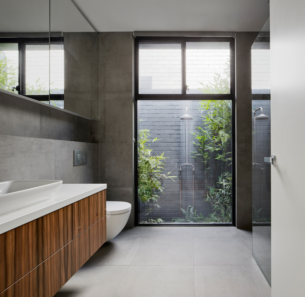 Photo of a mid-sized modern master wet room bathroom in Melbourne with furniture-like cabinets, medium wood cabinets, a freestanding tub, a wall-mount toilet, gray tile, porcelain tile, grey walls, porcelain floors, a drop-in sink, engineered quartz benchtops, grey floor, an open shower and white benchtops.