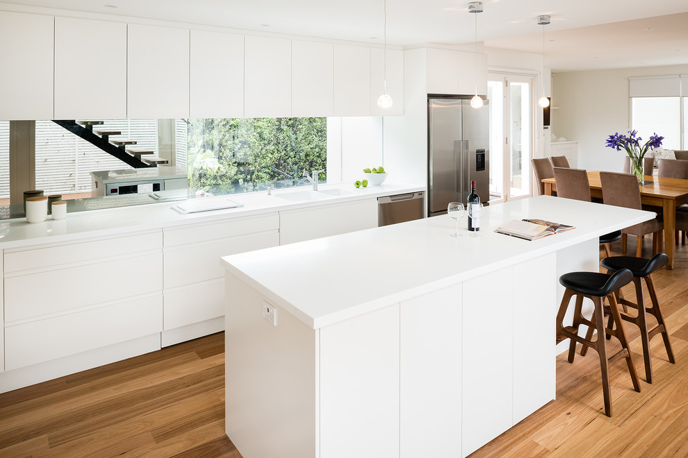 Photo of a mid-sized contemporary l-shaped eat-in kitchen in Melbourne with an integrated sink, flat-panel cabinets, white cabinets, solid surface benchtops, mirror splashback, stainless steel appliances, medium hardwood floors, with island, metallic splashback and white benchtop.