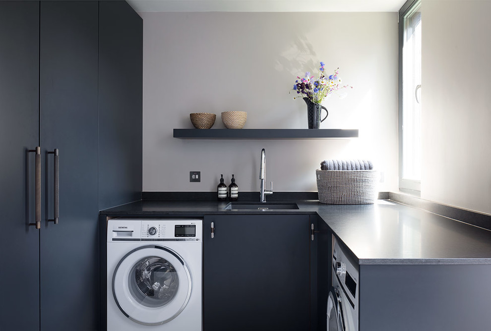 This is an example of a small contemporary u-shaped laundry room in London with an undermount sink, marble benchtops, light hardwood floors, flat-panel cabinets, black cabinets, grey walls, black benchtop and an integrated washer and dryer.