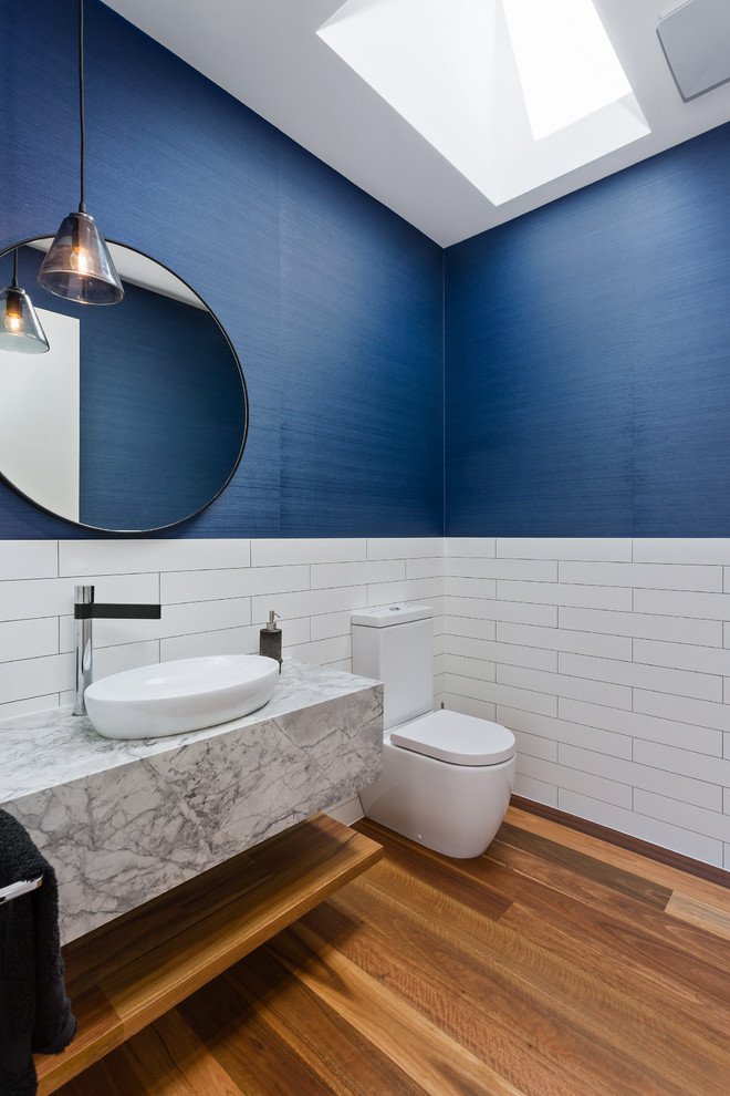 Inspiration for a mid-sized contemporary powder room in Melbourne with open cabinets, medium wood cabinets, a two-piece toilet, white tile, blue walls, medium hardwood floors, a vessel sink, brown floor, white benchtops and marble benchtops.