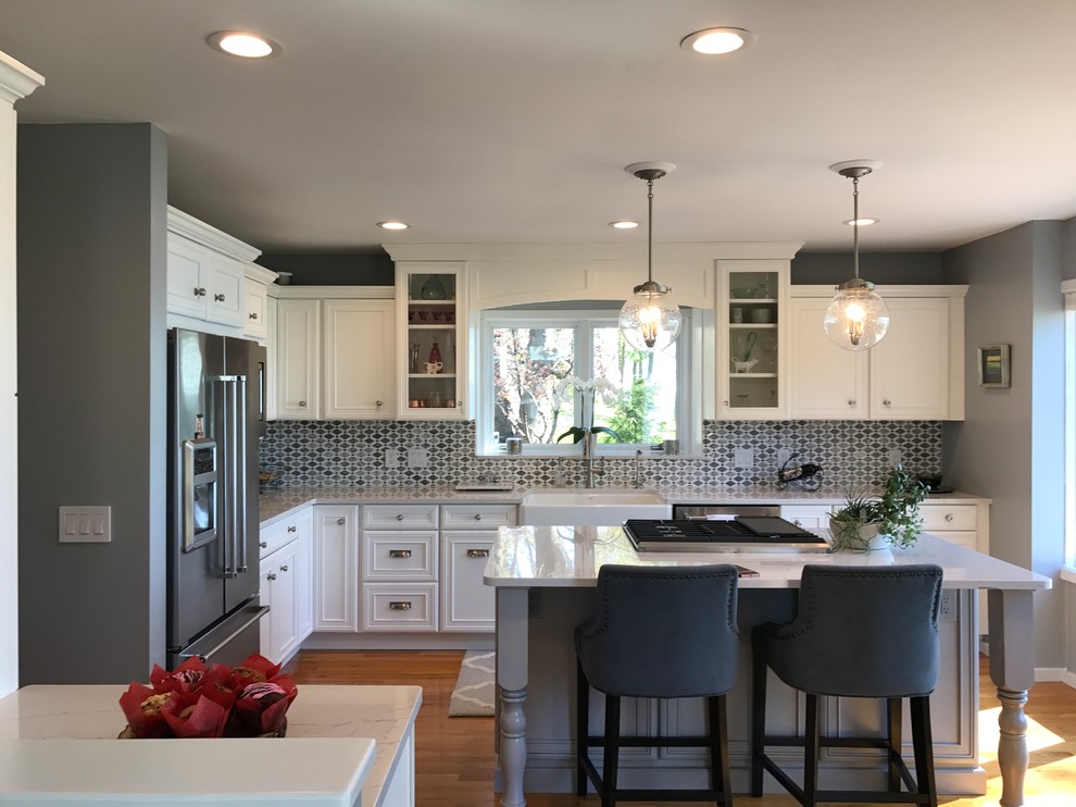 Example of a mid-sized transitional l-shaped medium tone wood floor and brown floor eat-in kitchen design with a farmhouse sink, recessed-panel cabinets, white cabinets, marble countertops, gray backsplash, mosaic tile backsplash, stainless steel appliances and an island