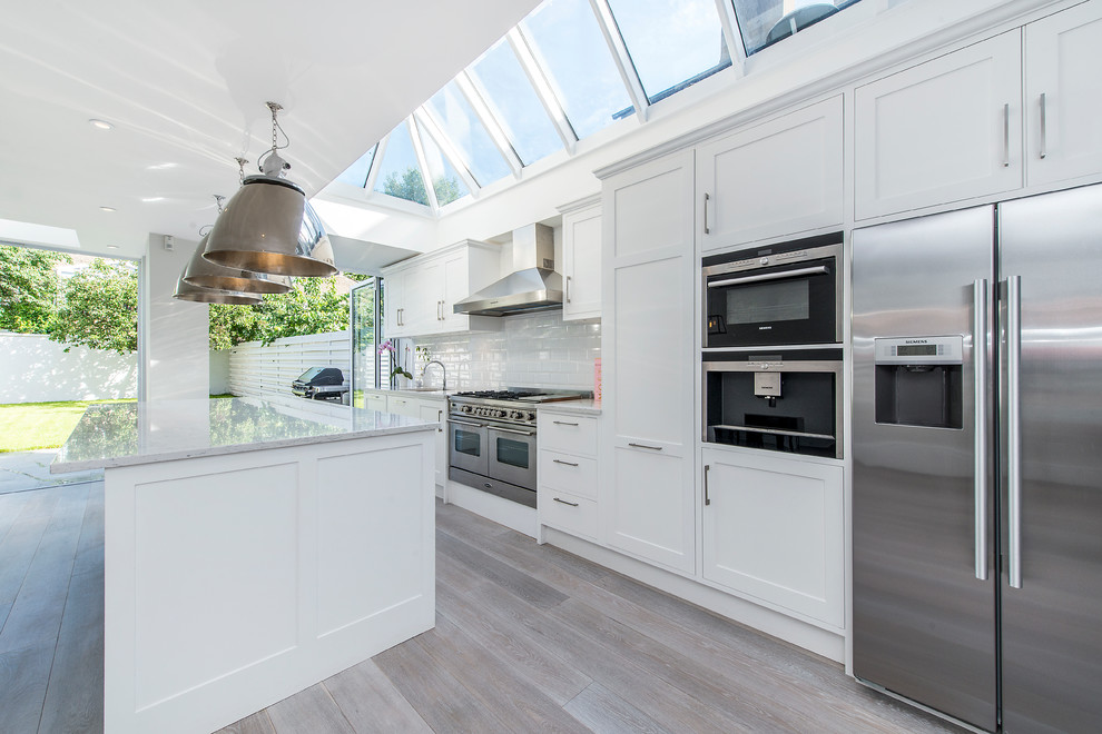 This is an example of a large contemporary single-wall open plan kitchen in London with a drop-in sink, recessed-panel cabinets, grey cabinets, marble benchtops, white splashback, subway tile splashback, stainless steel appliances, light hardwood floors and with island.