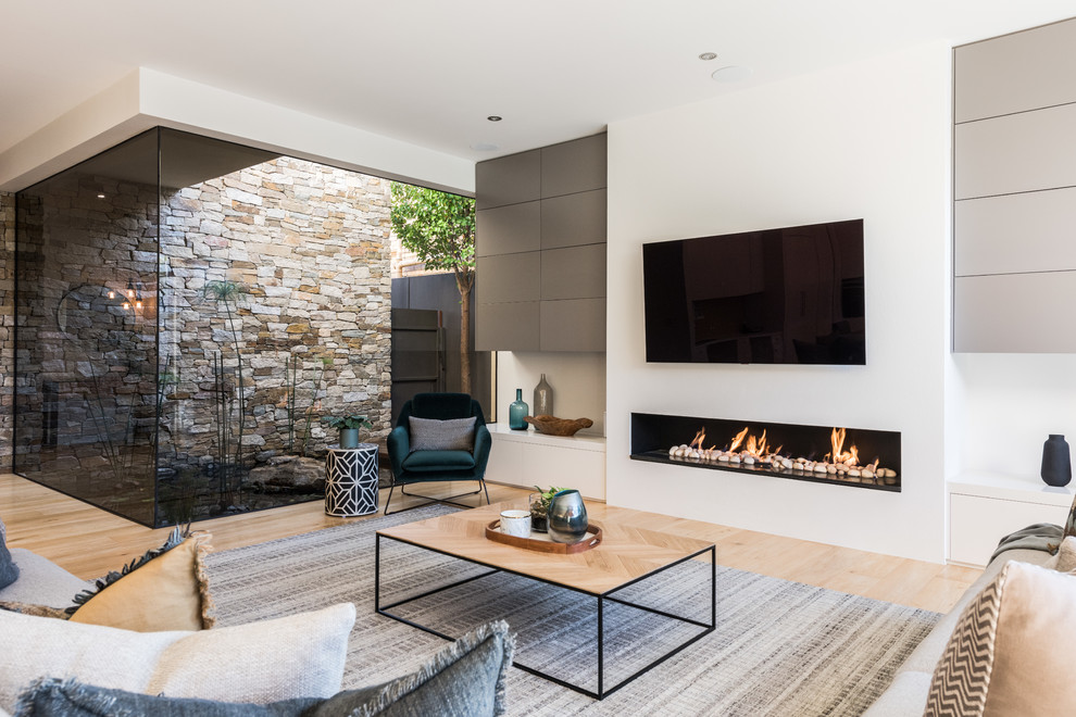 This is an example of a contemporary open concept living room in Melbourne with white walls, light hardwood floors, a ribbon fireplace, a wall-mounted tv and beige floor.
