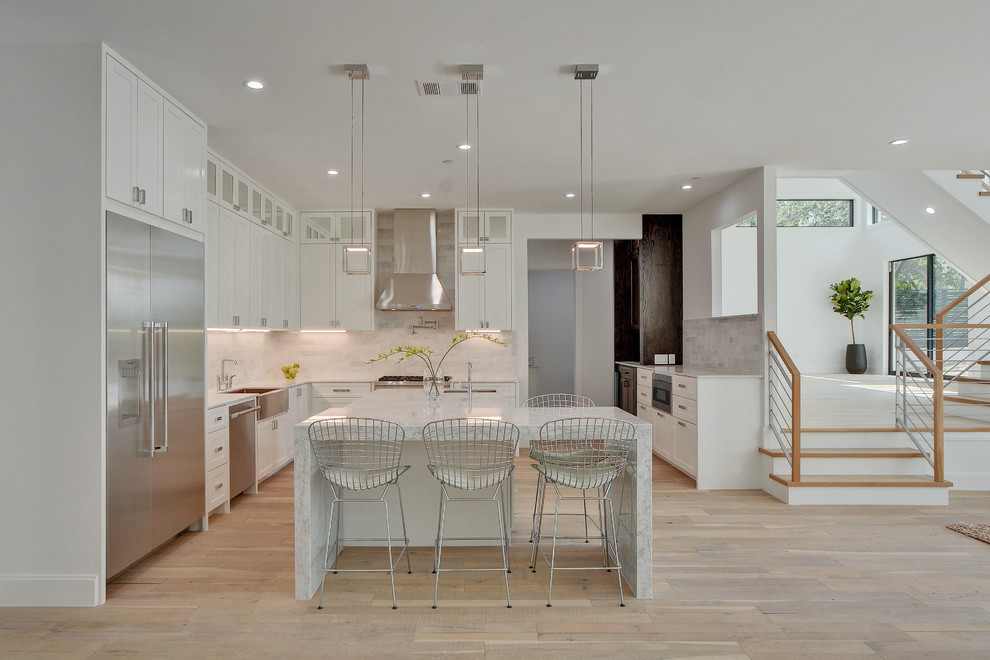 Design ideas for a large contemporary u-shaped open plan kitchen in Nashville with a farmhouse sink, shaker cabinets, white cabinets, white splashback, stainless steel appliances, light hardwood floors, with island, marble benchtops, stone tile splashback, beige floor and white benchtop.