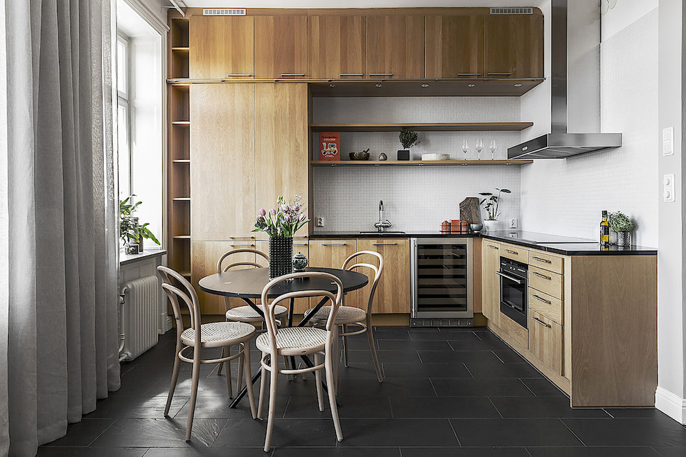 Mid-sized scandinavian l-shaped eat-in kitchen in Stockholm with flat-panel cabinets, light wood cabinets, no island, black benchtop, an undermount sink, white splashback, black appliances and black floor.