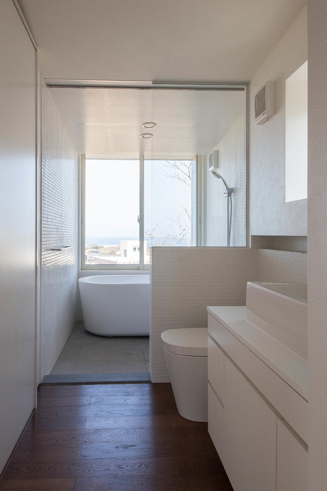 This is an example of a beach style bathroom in Other with flat-panel cabinets, white cabinets, a freestanding tub, a one-piece toilet, white walls, dark hardwood floors, a vessel sink, an open shower, a curbless shower, white tile and brown floor.