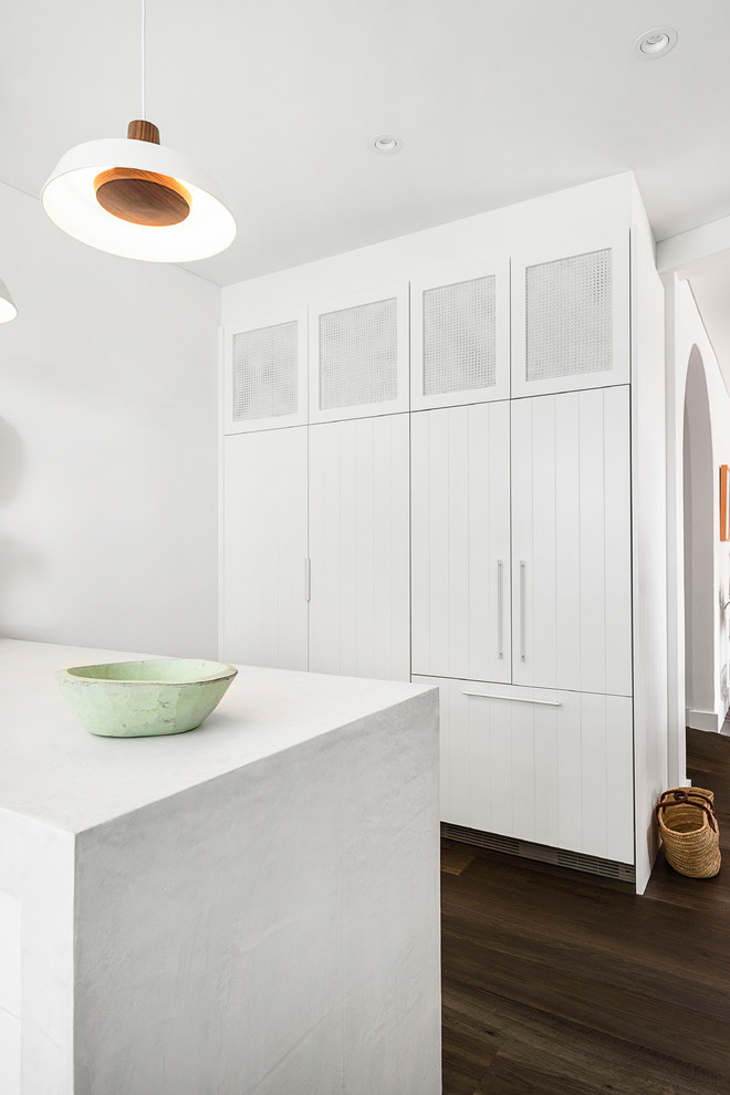 Photo of a mid-sized beach style l-shaped kitchen pantry in Sydney with an undermount sink, shaker cabinets, white cabinets, quartz benchtops, beige splashback, ceramic splashback, white appliances, dark hardwood floors, with island, brown floor and white benchtop.