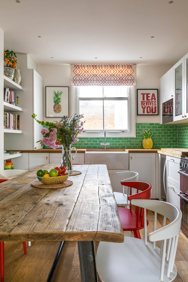 Design ideas for a mid-sized eclectic l-shaped eat-in kitchen in London with a farmhouse sink, shaker cabinets, white cabinets, wood benchtops, green splashback, ceramic splashback, stainless steel appliances, medium hardwood floors, no island, brown floor and brown benchtop.