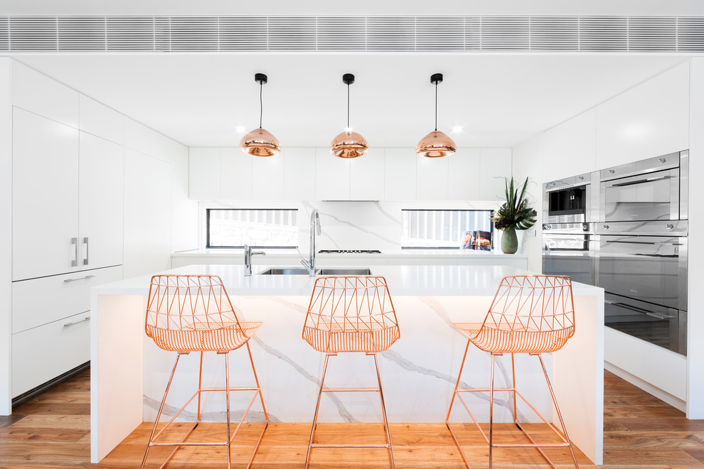 This is an example of a large contemporary u-shaped separate kitchen in Canberra - Queanbeyan with a double-bowl sink, flat-panel cabinets, white cabinets, quartz benchtops, white splashback, stone slab splashback, coloured appliances, medium hardwood floors, with island, white benchtop and brown floor.