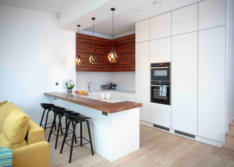 Photo of a mid-sized contemporary l-shaped eat-in kitchen in London with an integrated sink, light hardwood floors, with island, white benchtop, flat-panel cabinets, white cabinets, solid surface benchtops, white splashback, stone slab splashback, stainless steel appliances and brown floor.