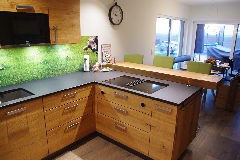 Design ideas for an expansive contemporary u-shaped open plan kitchen in Nuremberg with an undermount sink, flat-panel cabinets, medium wood cabinets, granite benchtops, green splashback, glass sheet splashback, black appliances, laminate floors, a peninsula and black benchtop.