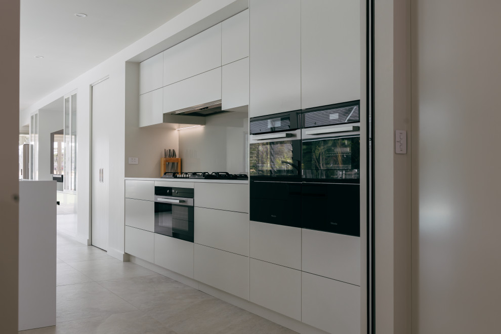 This is an example of a large modern galley open plan kitchen in Central Coast with an integrated sink, flat-panel cabinets, beige cabinets, engineered stone countertops, beige splashback, glass sheet splashback, black appliances, porcelain flooring, an island, beige floors and white worktops.