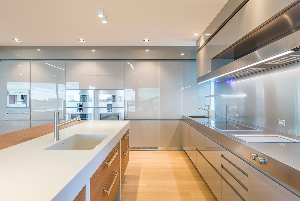 Photo of a mid-sized modern u-shaped eat-in kitchen in New York with an integrated sink, flat-panel cabinets, grey cabinets, stainless steel benchtops, metallic splashback, metal splashback, stainless steel appliances, light hardwood floors, multiple islands and beige floor.